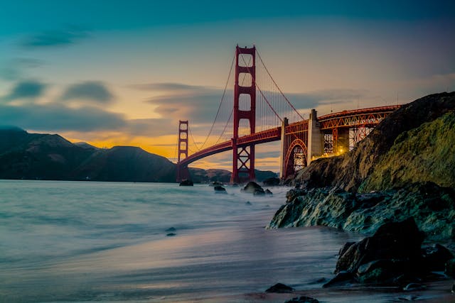 Golden Gate Bridge at sunset viewed from a rocky beach, with silky long-exposure waves crashing against the shore and the Marin Headlands in the background.