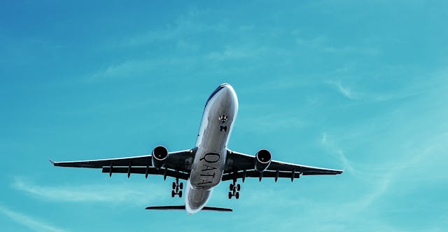Low-angle view of a Qatar Airways airliner flying overhead with landing gear deployed, set against a bright turquoise sky with wispy clouds