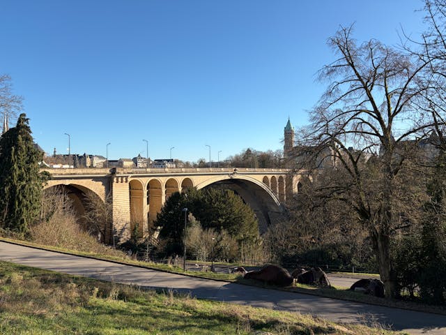 Le Pont Adolphe à Luxembourg, un imposant pont en arche de pierre enjambant une vallée boisée, sous un ciel bleu ensoleillé Le Pont Adolphe à Luxembourg, un imposant pont en arche de pierre enjambant une vallée boisée, sous un ciel bleu ensoleillé
